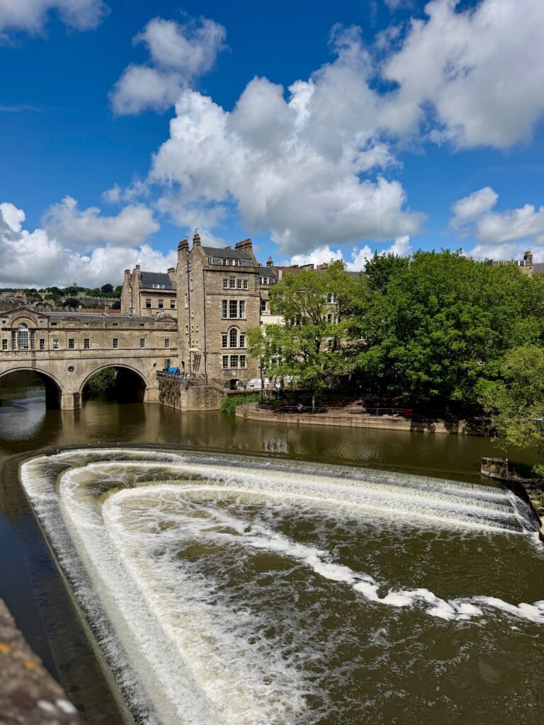 Pulteney Bridge spanning the River Avon, with its historic stone buildings and arches visible behind the curved horseshoe shaped weir. Water cascades over the weir beneath a bright blue sky filled with large white clouds.