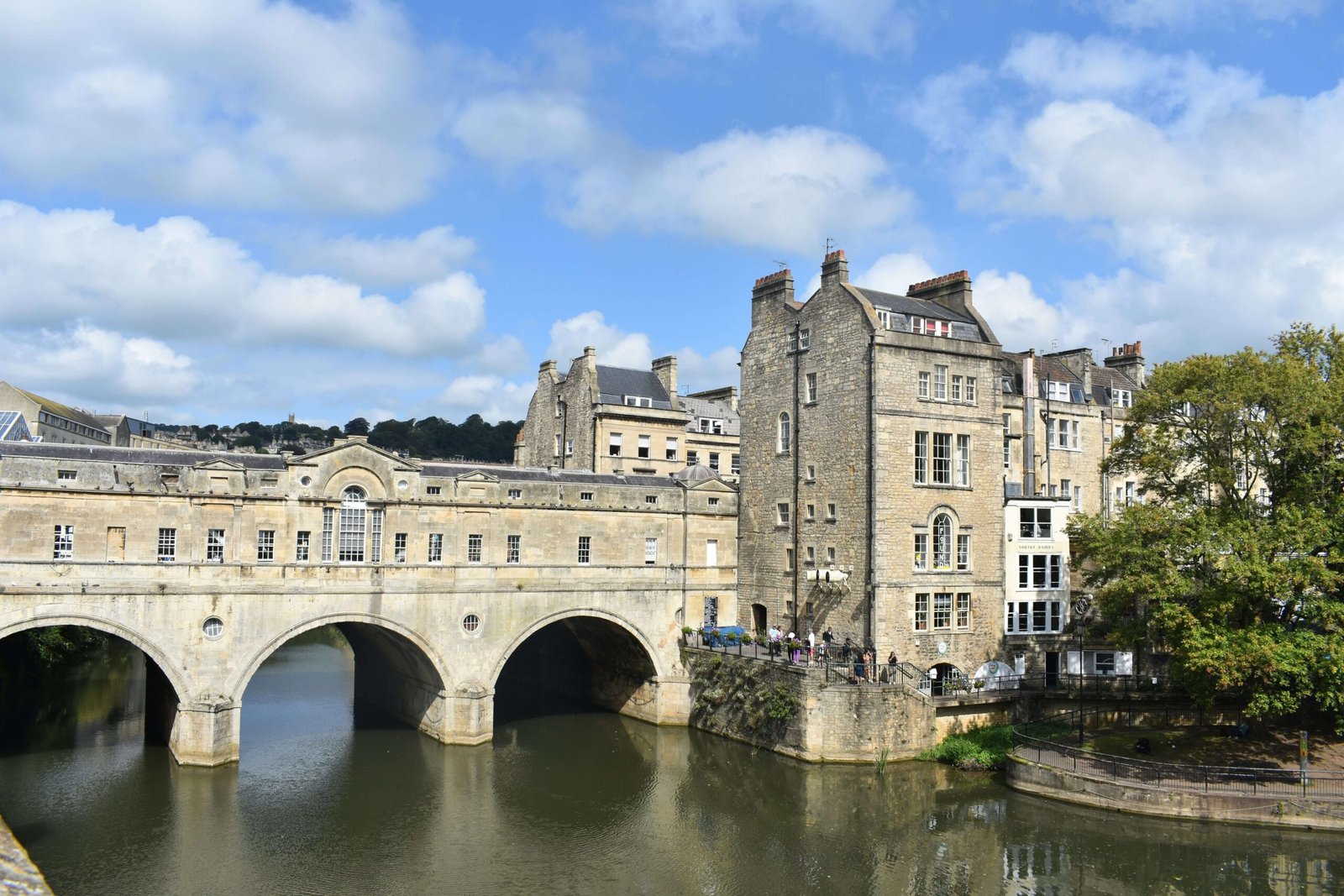 Pulteney Bridge spanning the River Avon in Bath, with its three stone arches and historic Georgian buildings reflected in the water below. Honey coloured Bath stone buildings line the riverbank beneath a bright blue sky dotted with white clouds.