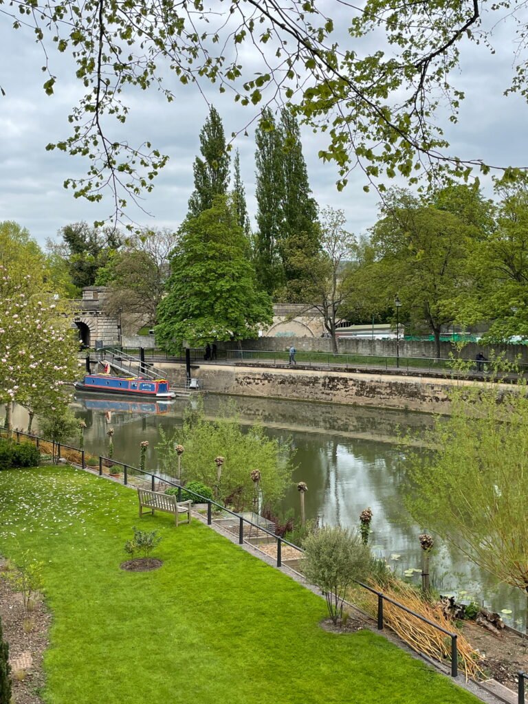A blue and red narrowboat moored along the River Avon beside a stone riverside walkway. A neatly maintained garden with a wooden bench and flowering trees overlooks the water, with mature green trees and historic stonework on the opposite bank.