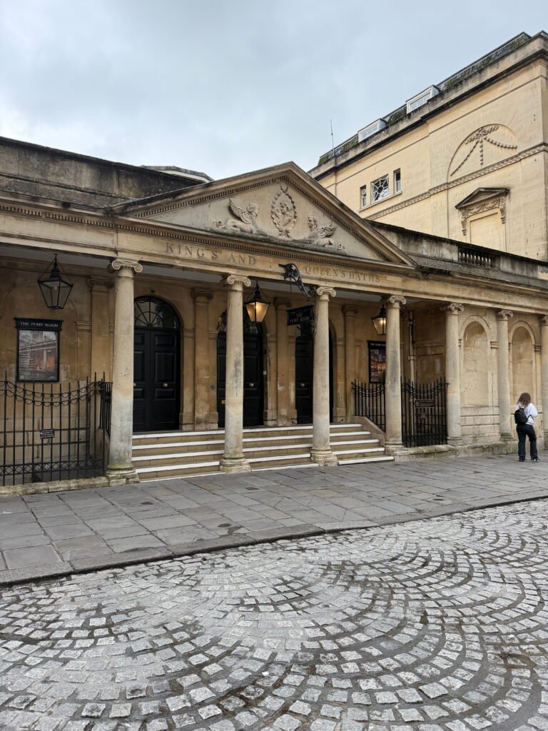 The historic entrance to the King's and Queen's Baths, with its classical stone columns, carved triangular pediment and black entrance doors. The words "KING'S AND QUEEN'S BATHS" are engraved above the columns, with signs for "THE PUMP ROOM" hanging beside the entrance.