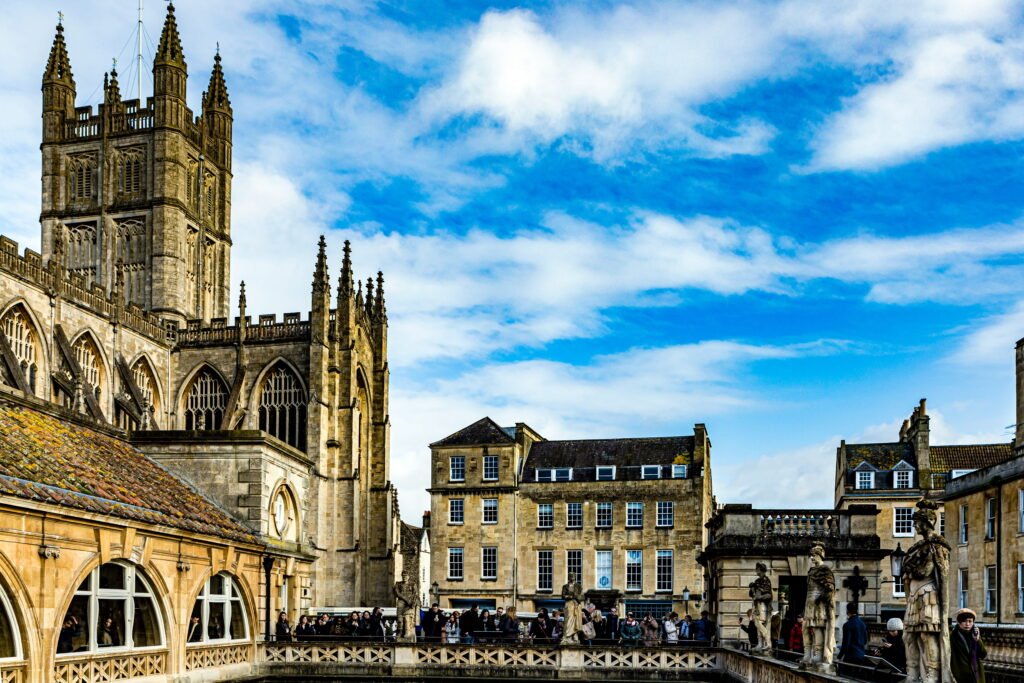 Bath Abbey towering above the Roman Baths in Bath, with intricate Gothic stonework, pointed arches and ornate spires against a bright blue sky with white clouds. Visitors gather around the historic Roman Baths terrace, where classical stone statues overlook the ancient bathing complex surrounded by honey coloured Bath stone buildings.