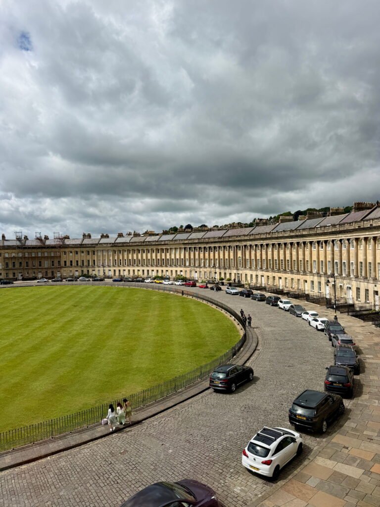 A panoramic view of the Royal Crescent, showing its sweeping curve of Georgian terraced houses with tall stone columns overlooking a vast green lawn. Cars line the cobbled road in front of the historic buildings beneath a dramatic cloudy sky.