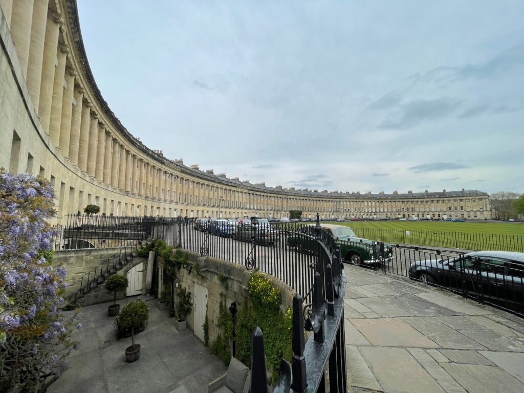 A private courtyard garden below street level at the Royal Crescent, with purple wisteria climbing the Georgian stone façade. Black wrought iron railings separate the garden from the curved row of historic terraced houses and parked cars above.