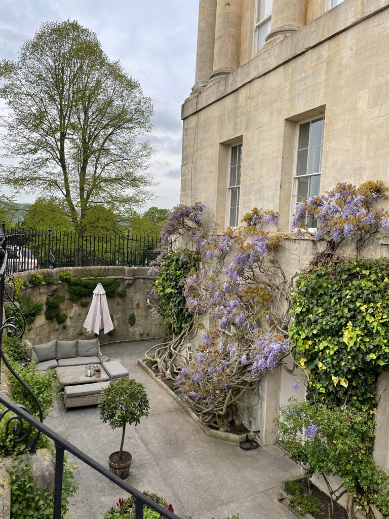 A secluded courtyard garden beside a Georgian townhouse, with flowering purple wisteria climbing the honey coloured stone walls. A grey outdoor sofa, matching footstool and closed parasol create a small seating area surrounded by greenery.