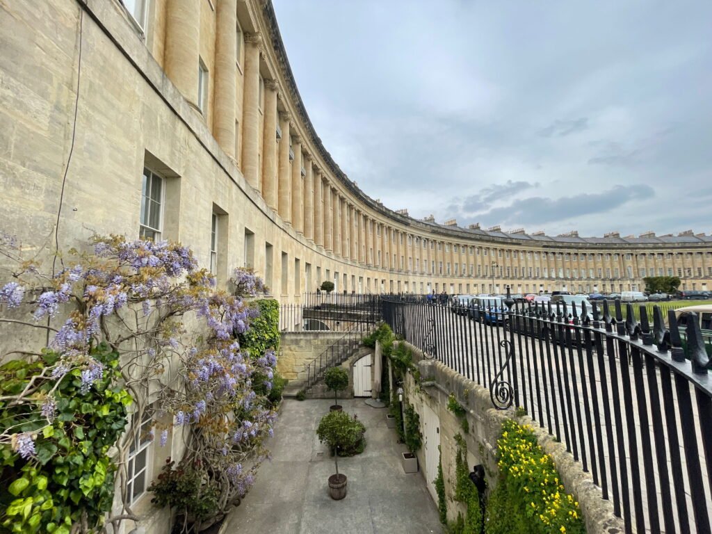 A private courtyard garden below street level at the Royal Crescent, with purple wisteria climbing the Georgian stone façade. Black wrought iron railings separate the garden from the curved row of historic terraced houses and parked cars above.