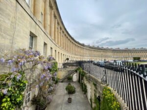 A private courtyard garden below street level at the Royal Crescent, with purple wisteria climbing the Georgian stone façade. Black wrought iron railings separate the garden from the curved row of historic terraced houses and parked cars above.