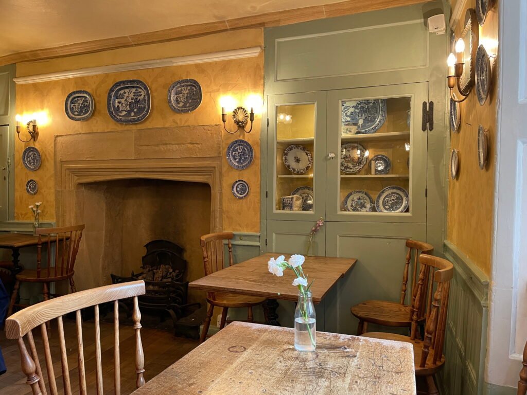 A cosy traditional café with yellow patterned walls, sage green woodwork and rustic wooden tables decorated with small vases of flowers. Blue and white decorative plates surround an old stone fireplace, with more porcelain displayed inside a glass fronted cupboard.