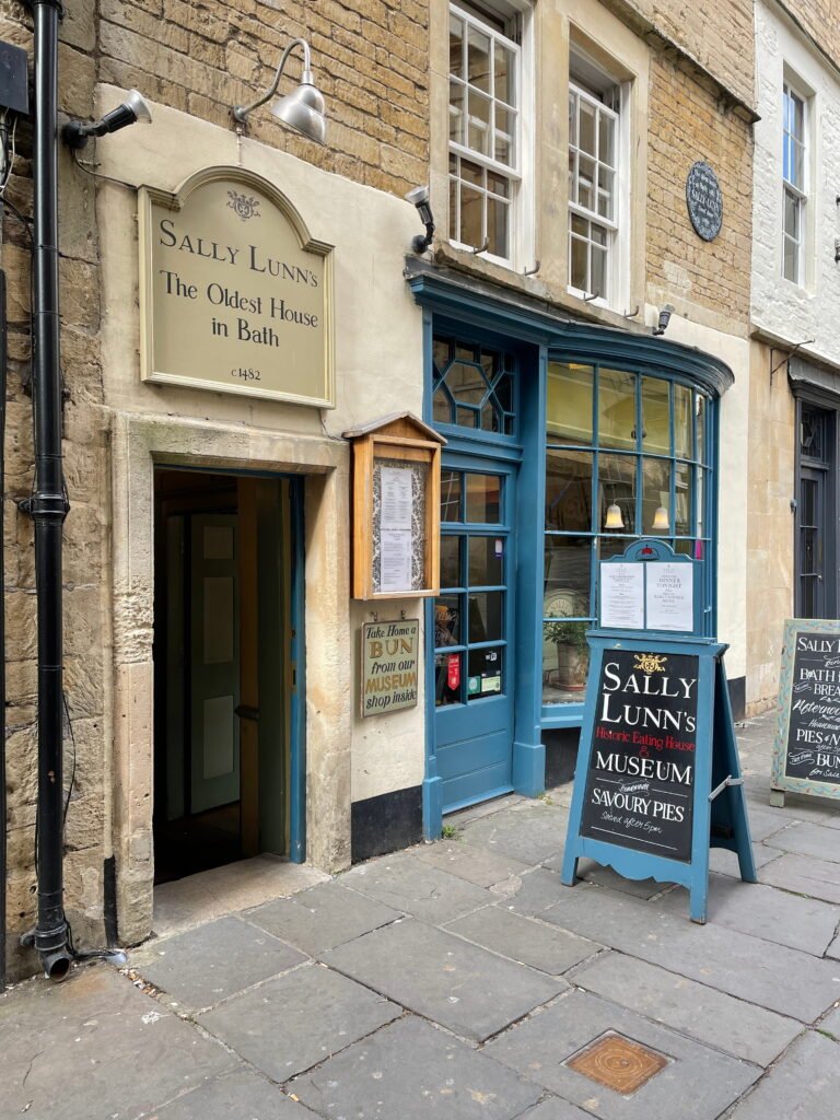 The exterior of Sally Lunn’s Historic Eating House and Museum in Bath features a blue shopfront set into an old stone building. A cream sign above the entrance reads "Sally Lunn’s. The Oldest House in Bath. c1482" while a pavement chalkboard advertises savoury pies and the historic eating house and museum.