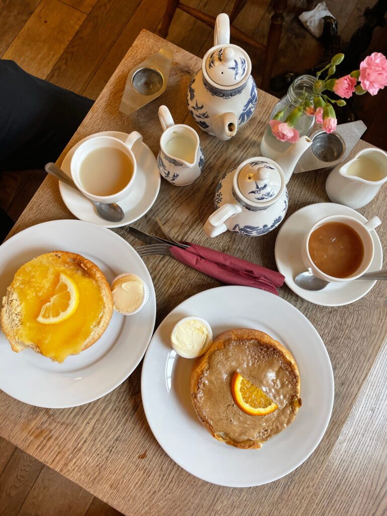 Two pastries topped with glossy icing and orange slices served with small portions of clotted cream on white plates. Two cups of tea, blue and white porcelain teapots and a vase of pink flowers complete the afternoon tea setting on a rustic wooden table.