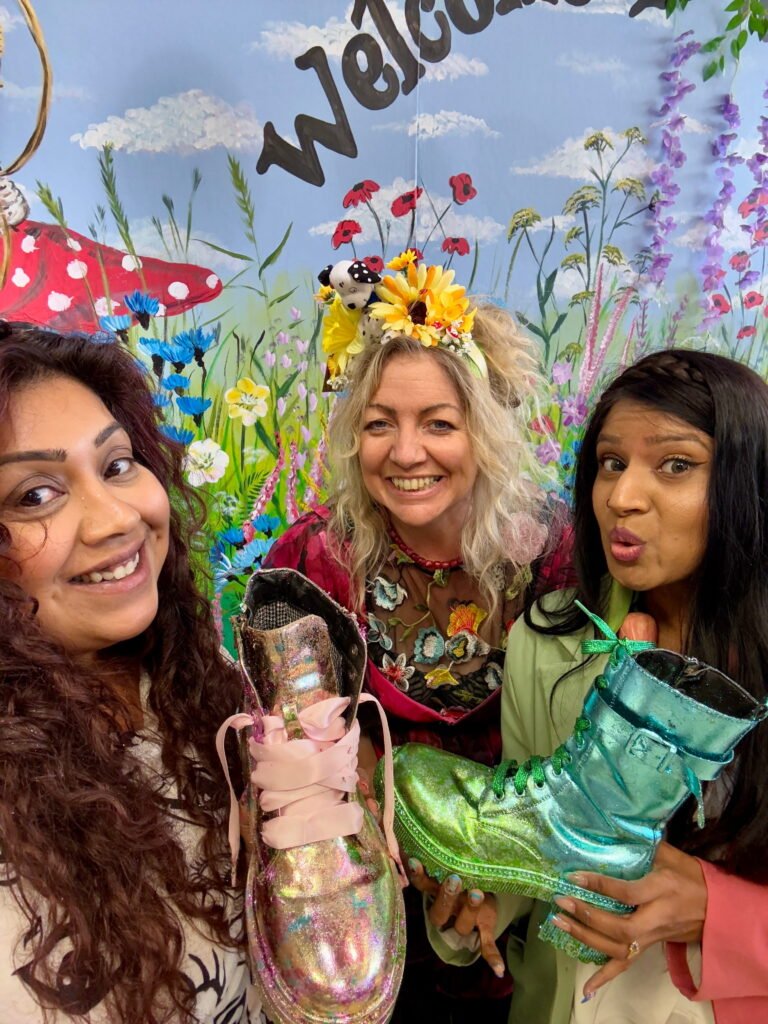 Three smiling women pose together in front of a colourful painted flower wall while holding oversized glittery pink, green and turquoise boots. The mural behind them reads "Welcome" above a meadow of flowers.