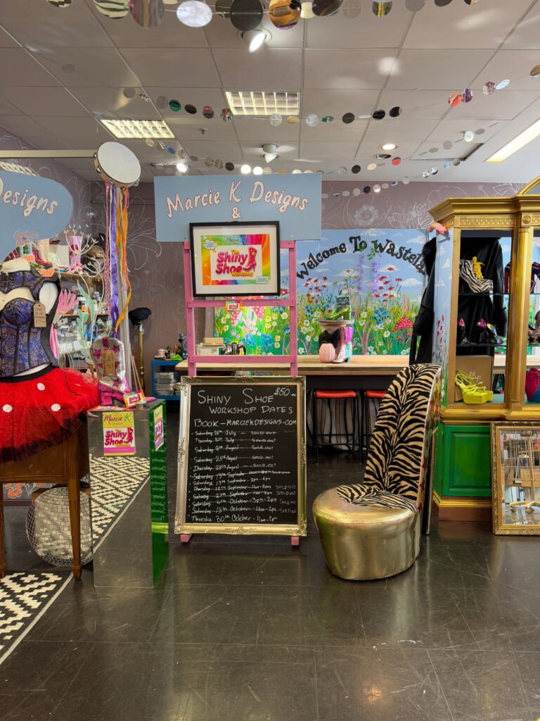 Colourful interior of Marcie K Designs with whimsical displays, a zebra print chair and hand painted floral decor. Signs read "Marcie K Designs" and "Welcome To Wasteland" with a chalkboard listing Shiny Shoe Workshop dates.