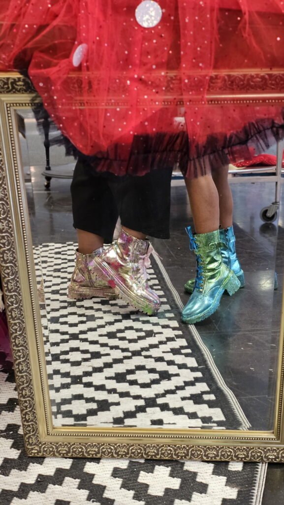 Two pairs of colourful customised boots reflected in an ornate floor mirror. One pair is metallic pink and gold while the other is metallic green and blue beneath the hem of a red tulle skirt.