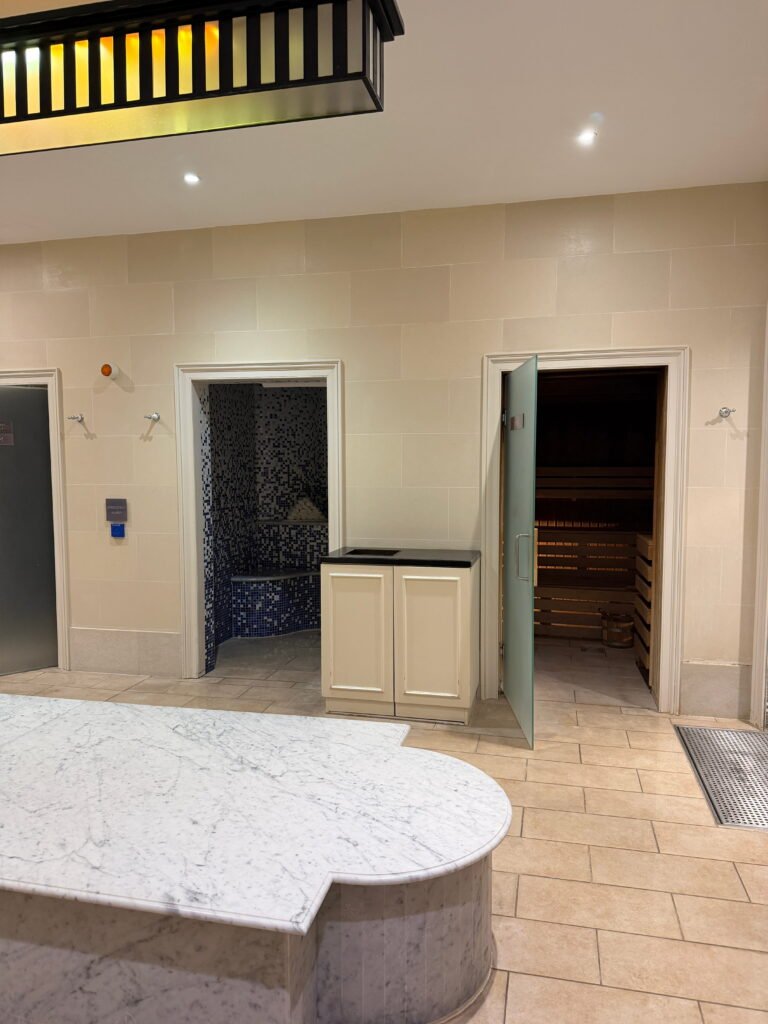 The spa at The Gainsborough Bath Spa features a pale tiled interior with a large white marble surface in the foreground. An open doorway reveals a blue mosaic tiled steam room while another leads into a wooden sauna with tiered benches.