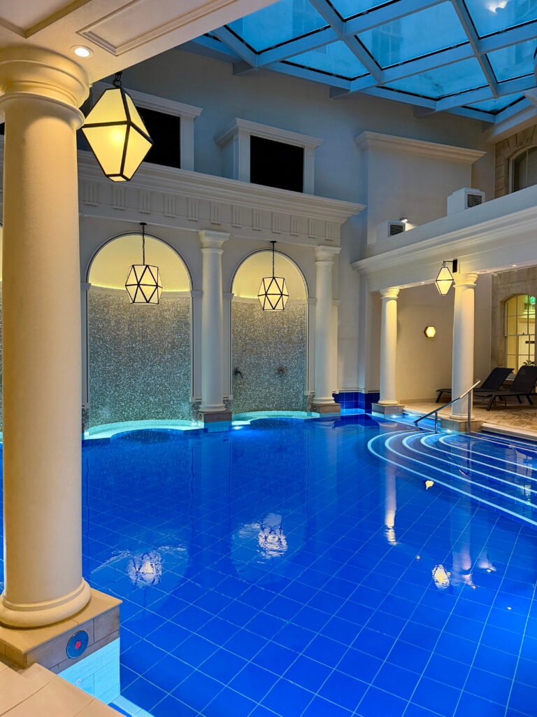 The indoor spa pool at The Gainsborough Bath Spa has vivid blue tiles and curved underwater steps leading into the water. White columns, illuminated mosaic walls and geometric pendant lanterns surround the pool beneath a large glass ceiling.