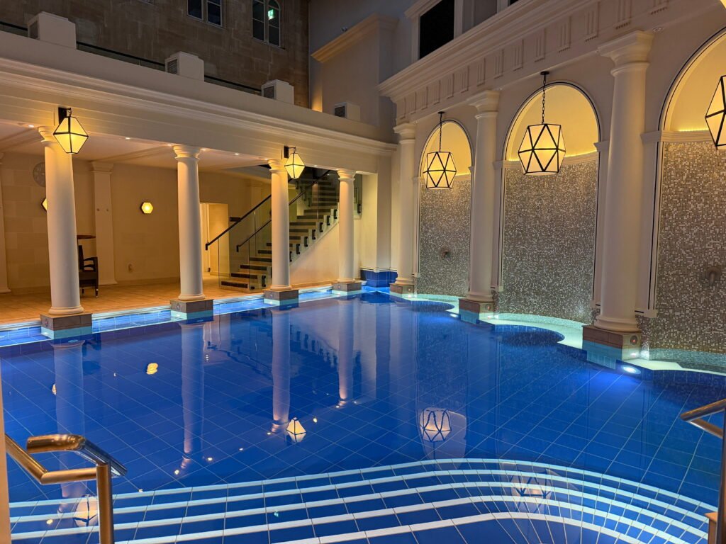 A wide view of the indoor spa pool at The Gainsborough Bath Spa showing blue tiled water, curved entry steps and white classical columns. Warm geometric pendant lights hang in front of arched mosaic walls while a staircase leads up from the pool area.