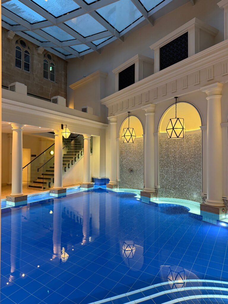 The Gainsborough Bath Spa indoor pool sits beneath a large glass roof framed by pale stone walls and white classical columns. Blue tiled water reflects the warm pendant lights hanging in front of tall arched mosaic walls.