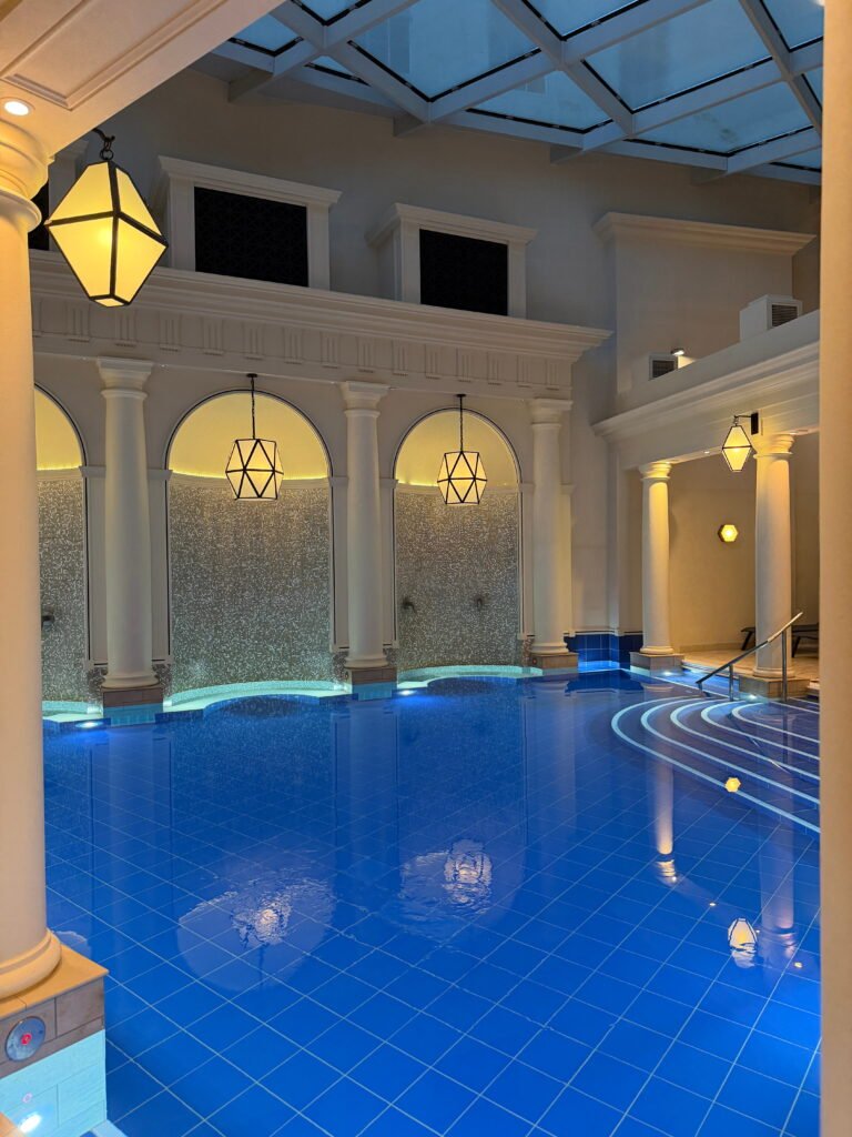 The indoor thermal spa pool at The Gainsborough Bath Spa features deep blue tiles, softly illuminated curved steps and tall white columns. Geometric lanterns hang above the water while mosaic tiled walls and a glass ceiling complete the elegant spa interior.