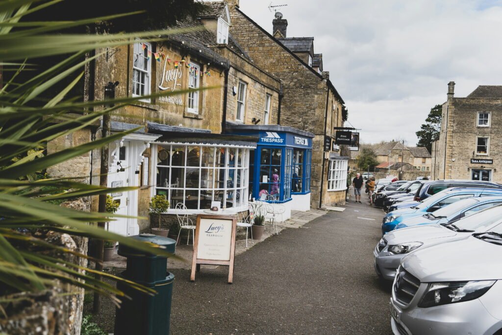Village street lined with stone shops and parked cars outside Lucys Tearoom. Storefront signs read "Lucys TEAROOM" "TRESPASS" "FINE PAINTINGS" and "TARA ANTIQUES" while a pavement sign reads "Lucys TEAROOM Quality tea and coffee Breakfasts Light lunches."