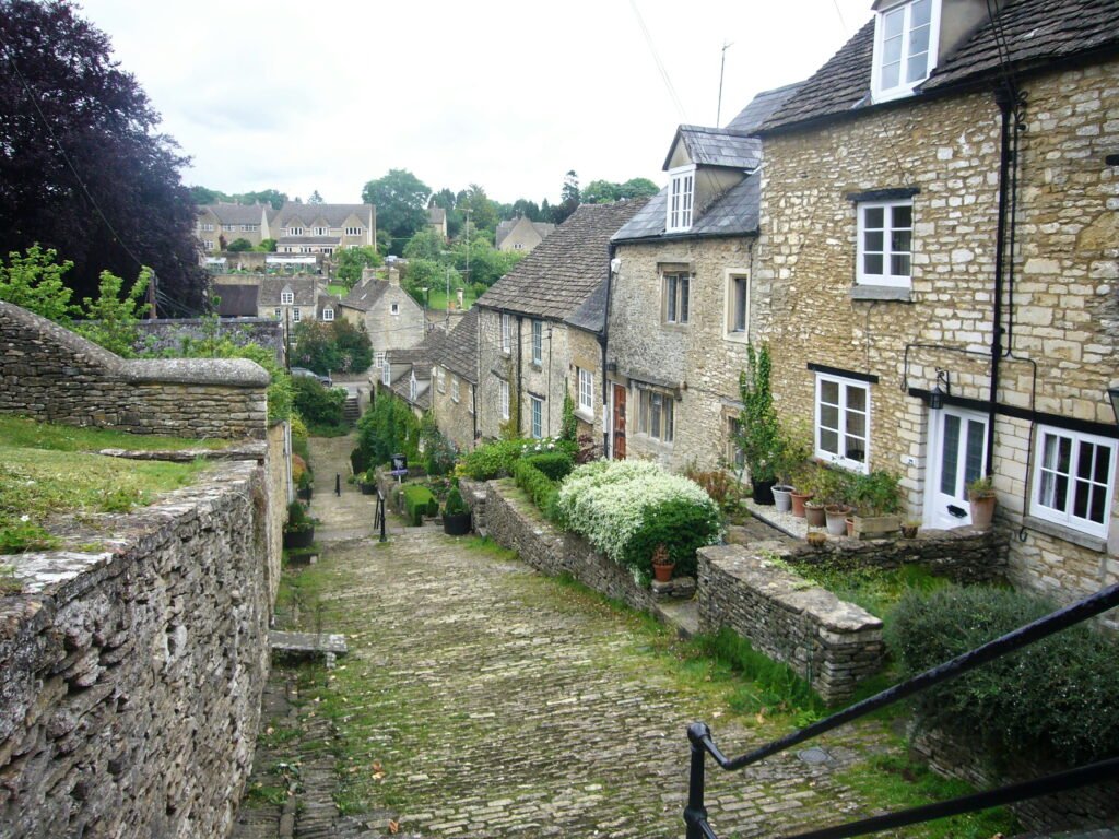 Steep stone lane descending between traditional Cotswold cottages and low stone garden walls. Potted plants shrubs and climbing greenery decorate the honey colored stone homes along the narrow path.