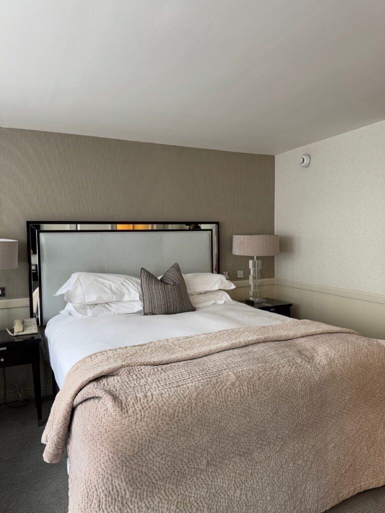 Neutral hotel bedroom with a large bed dressed in crisp white bedding, a textured beige bedspread and striped cushion. Matching bedside lamps and soft patterned walls give the room a calm understated look.