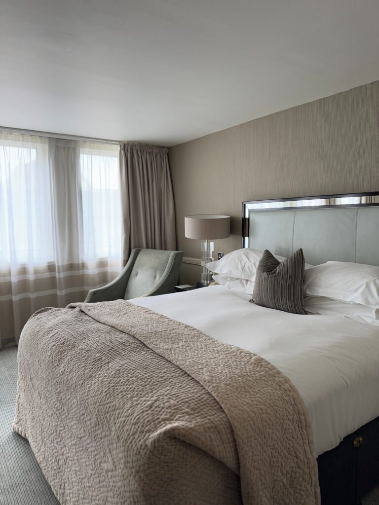 Large hotel bed beside a bright window with sheer curtains and a pale upholstered armchair. White bedding, a beige textured bedspread and neutral furnishings create a light comfortable room.