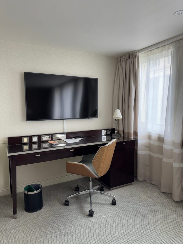 Hotel room workspace with a large wall mounted television above a dark desk and wheeled chair. A tall window with sheer curtains brings natural light into the neutral room.