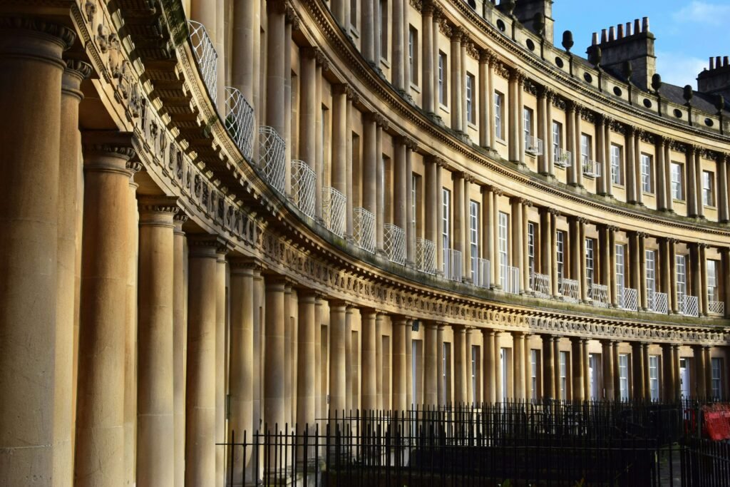 The curved Georgian architecture of The Circus in Bath, featuring rows of tall classical columns, ornate stone carvings and sash windows across three storeys. Warm sunlight highlights the honey coloured Bath stone façade, with black wrought iron railings running along the front of the historic townhouses.