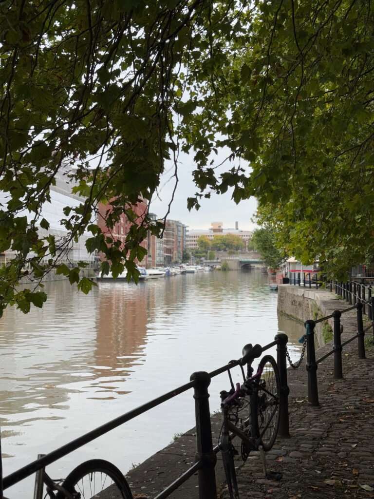 Quiet Bristol waterside path shaded by leafy trees with a bicycle leaning against black railings. Boats and city buildings are visible across the calm harbour.
