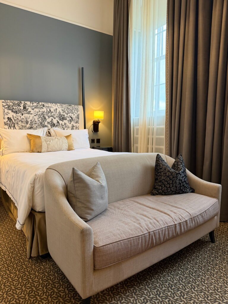 A cream upholstered sofa with grey decorative cushions sits at the foot of a large white bed in a bedroom at The Gainsborough Bath Spa. Floor length curtains frame a tall window while a gold accent cushion and patterned headboard add detail to the room.