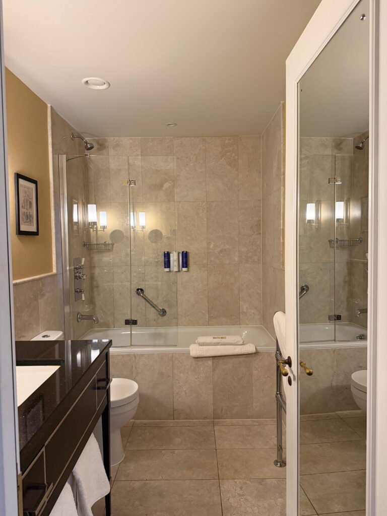 A spacious bathroom at The Gainsborough Bath Spa features beige marble effect tiles, a bathtub with a glass shower screen and a dark vanity unit. Folded white towels rest on the edge of the bath beneath wall mounted toiletries and a large mirror reflects the shower area.