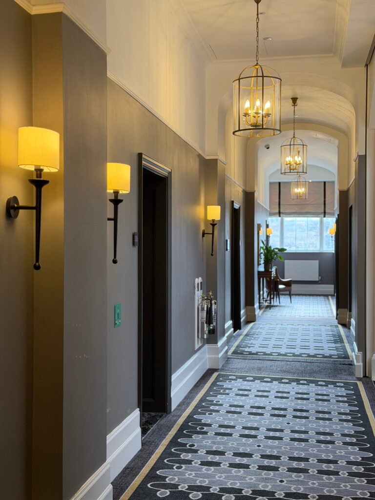 A long corridor at The Gainsborough Bath Spa features grey walls, white arched ceilings and a patterned grey carpet. Warm wall sconces and gold framed pendant lanterns illuminate the hallway leading towards a bright window and seating area.