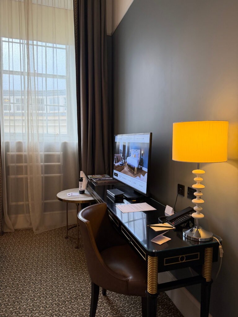 A black writing desk with gold detailing sits beside a tall curtained window in a bedroom at The Gainsborough Bath Spa. A television, hotel telephone, decorative table lamp and brown leather chair create a dedicated workspace within the room.