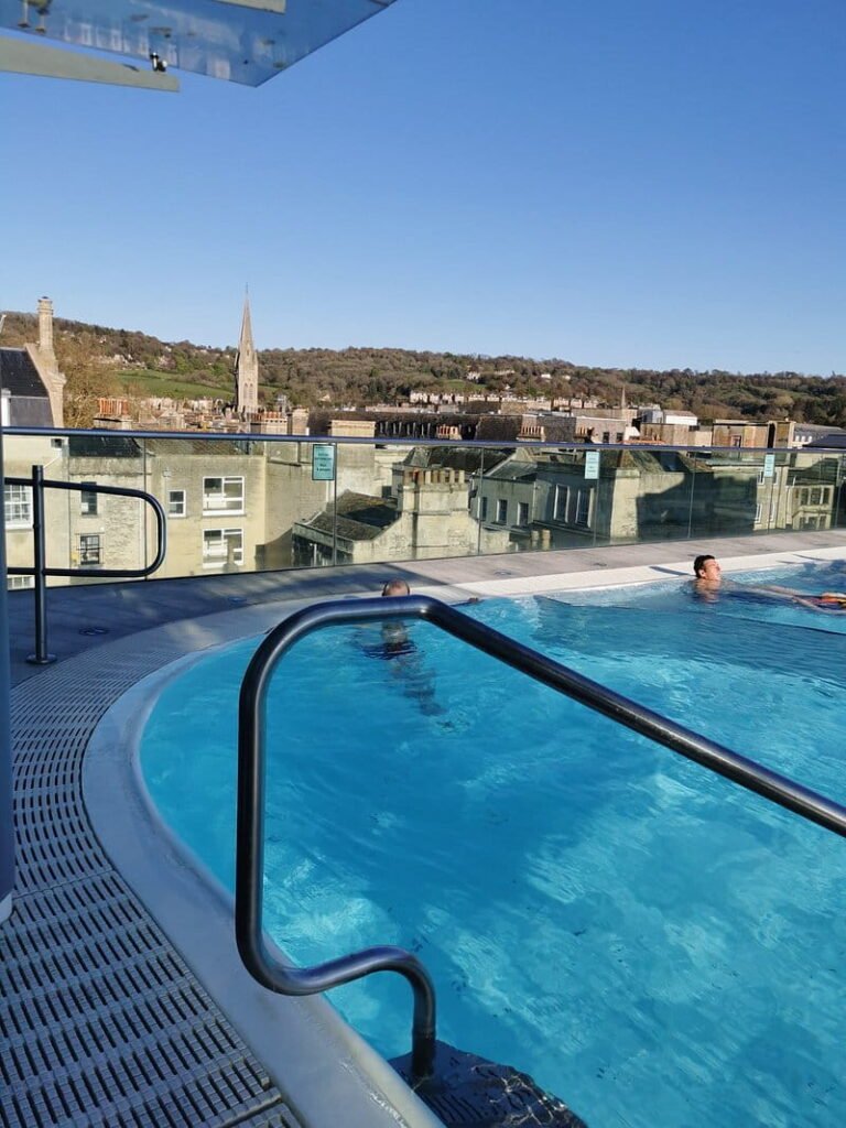 An open air rooftop swimming pool at Thermae Bath Spa, with bathers enjoying the warm blue water overlooking Bath's historic rooftops and a church spire. A glass balustrade provides uninterrupted views of the surrounding city and green hills beneath a clear blue sky.