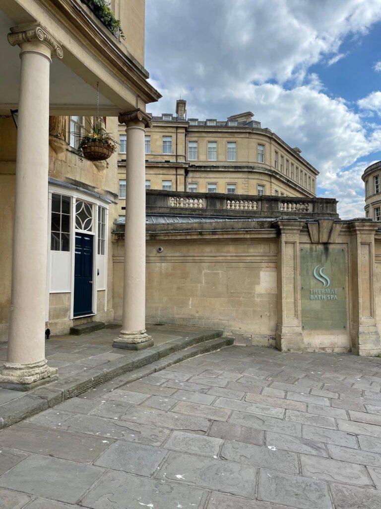 The exterior of Thermae Bath Spa, with a glass sign reading "THERMAE BATH SPA" mounted on a honey coloured stone wall. Classical columns, a dark blue entrance door and a hanging flower basket frame the paved courtyard beneath a partly cloudy sky.