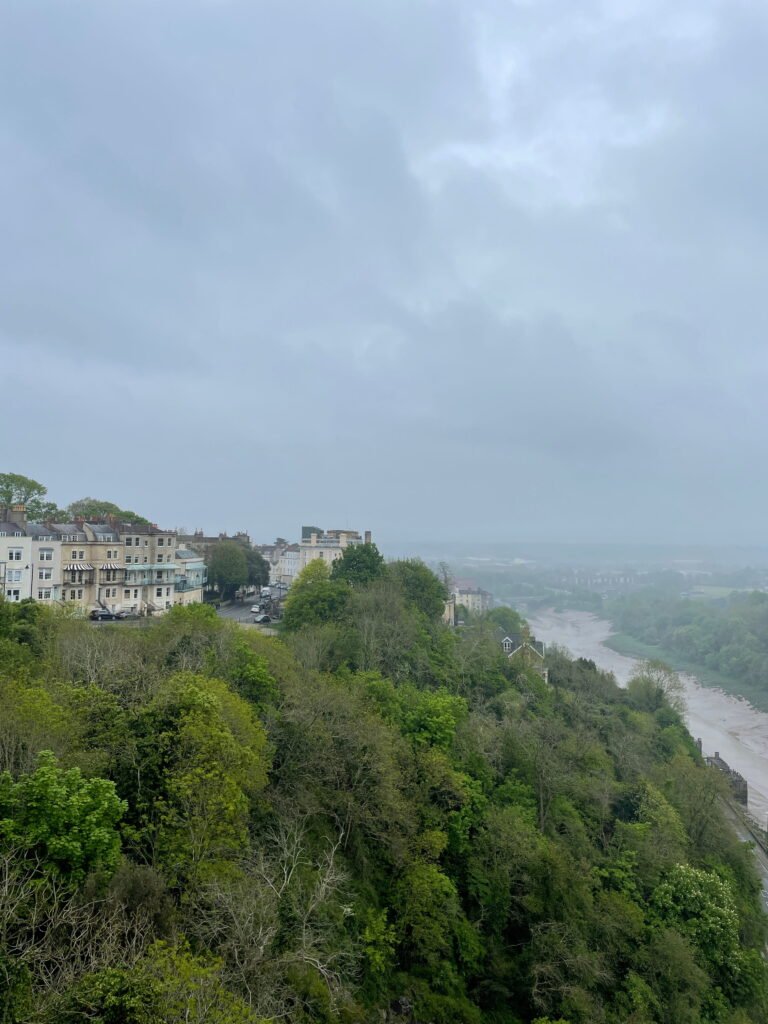Colourful houses sit high above the River Avon on a densely wooded hillside in Bristol. Mist softens the distant view along the gorge and river below.