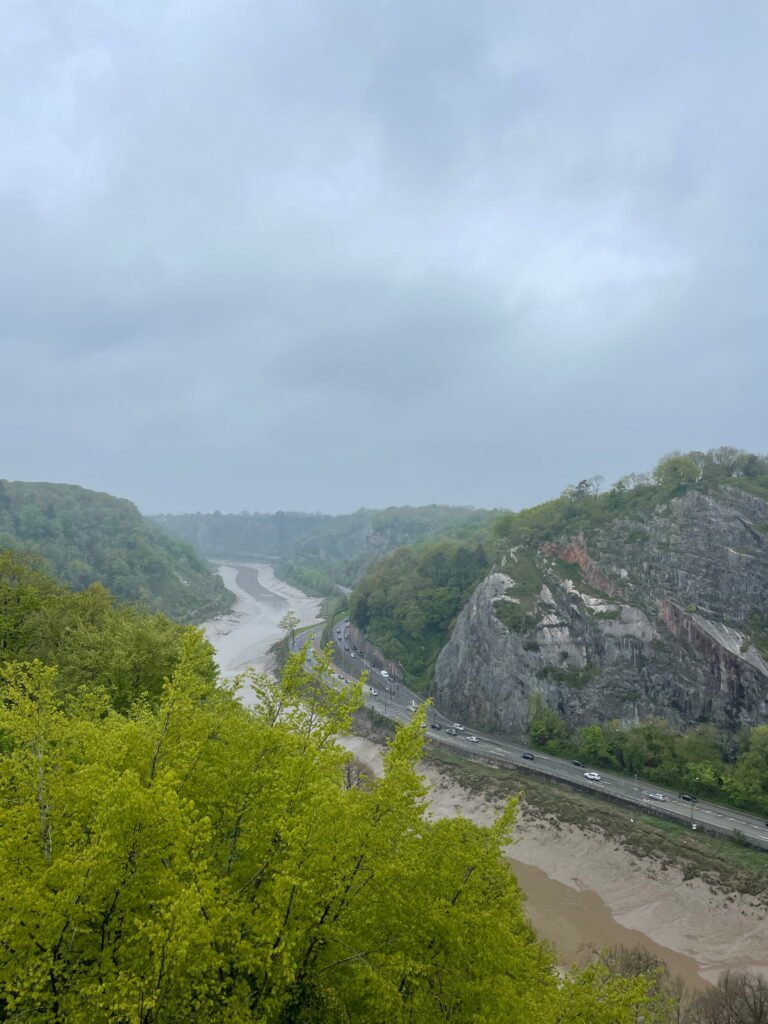 A sweeping view along the Avon Gorge with the River Avon winding between wooded cliffs. A road curves around the base of the rocky gorge beneath a cloudy grey sky.