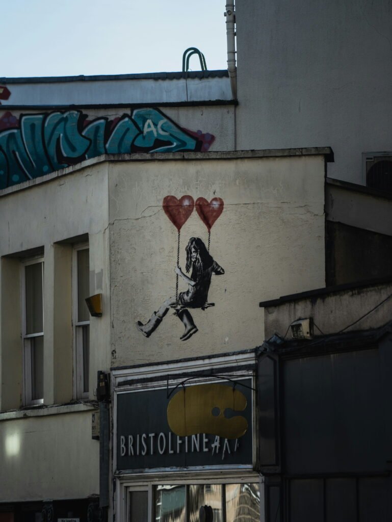 Street art of a girl sitting on a swing suspended from two red heart shaped balloons on a wall in Bristol. The artwork appears above a shop sign reading "BRISTOL FINE ART".