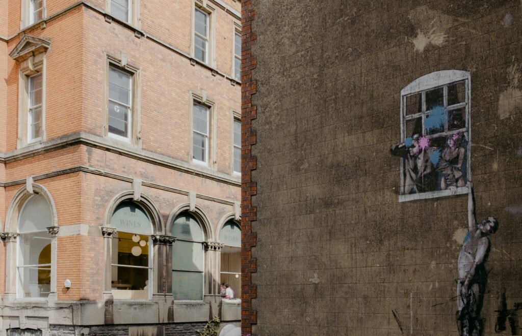 Street art painted on a weathered wall in Bristol shows figures leaning from a painted window while another figure reaches upwards from below. Splashes of blue and pink paint cover the faces of the figures.