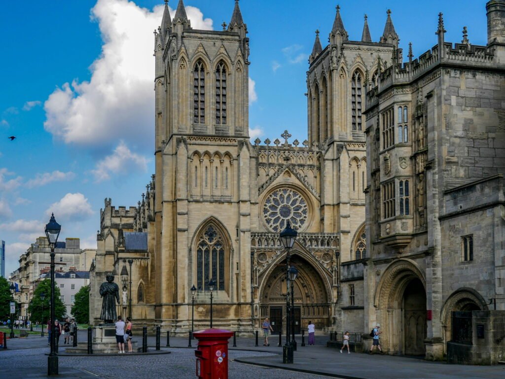 Bristol Cathedral with its twin Gothic towers and ornate stone entrance beneath a bright blue sky. People walk through College Green while a red post box and statue stand in front of the cathedral.