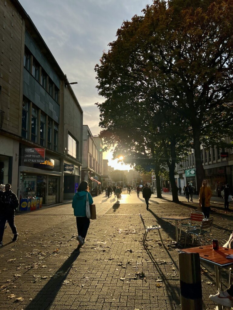 People walk along a pedestrianised shopping street in central Bristol as low evening sunlight shines through large trees. Long shadows stretch across the pavement and outdoor tables beneath the golden light.