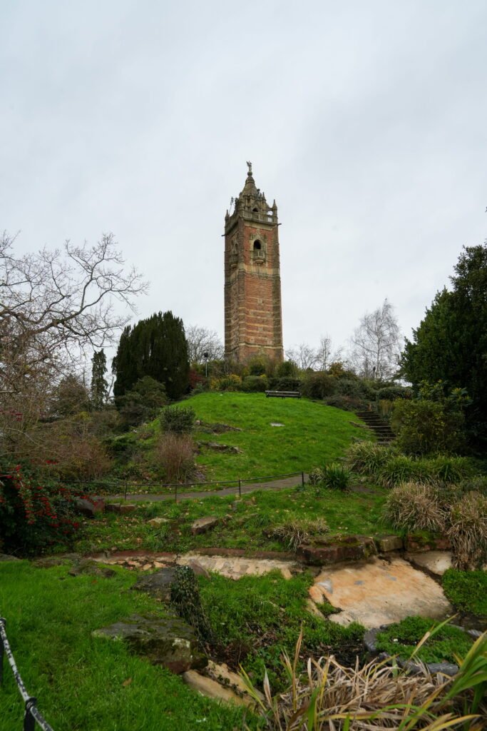 Cabot Tower rises above the lawns and gardens of Brandon Hill in Bristol. The tall red stone tower stands among trees and shrubs beneath an overcast sky.