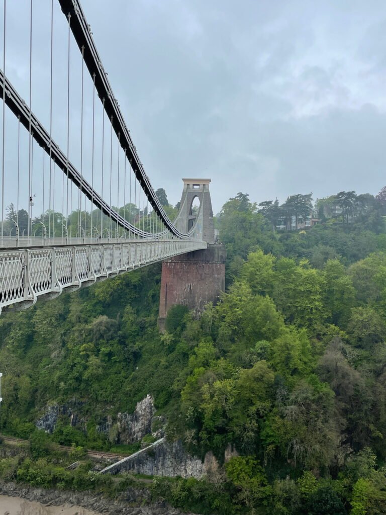 Clifton Suspension Bridge extends towards its stone tower above the densely wooded Avon Gorge. The pale metal bridge and suspension cables stand out against the green hillside and cloudy sky.
