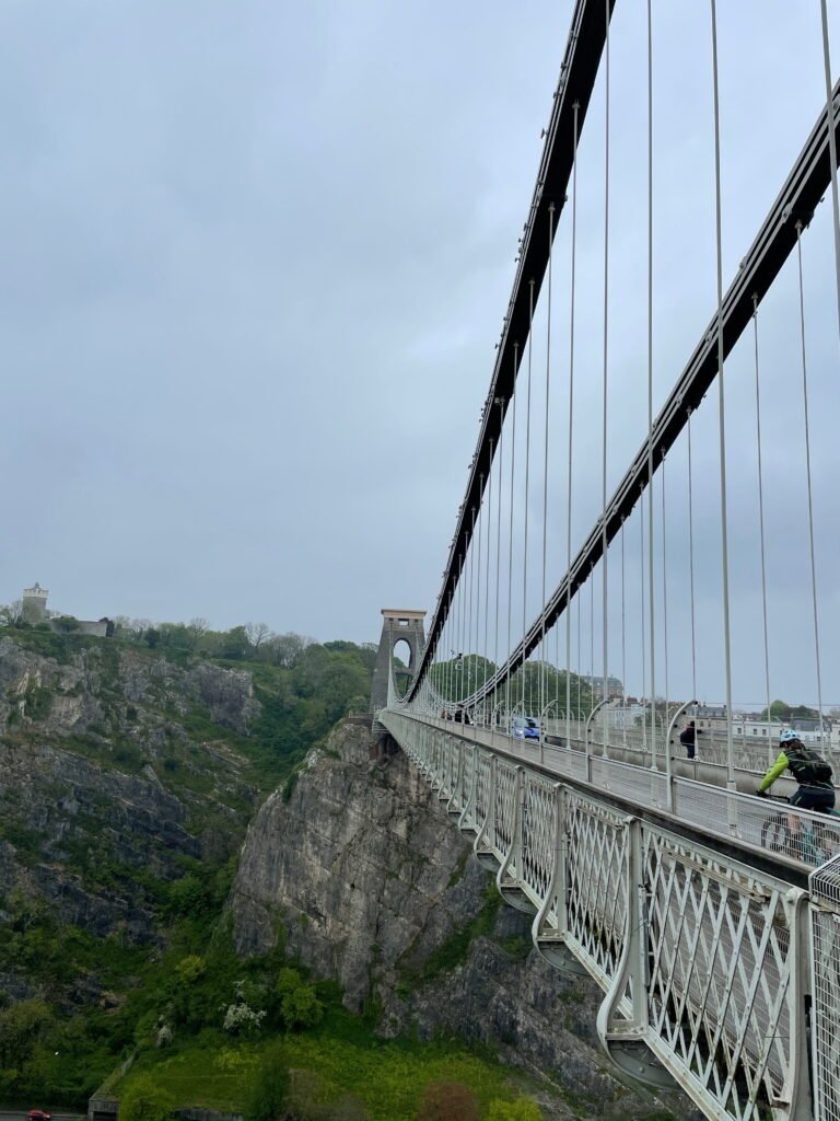 Clifton Suspension Bridge stretches across the rocky Avon Gorge in Bristol beneath an overcast sky. Cyclists and other travellers cross the bridge while the suspension cables rise overhead.