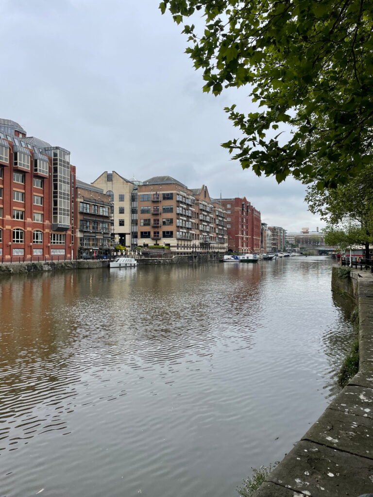 Boats moored along Bristols Floating Harbour beside red brick and modern waterfront buildings. Leafy branches frame the calm water on an overcast day.
