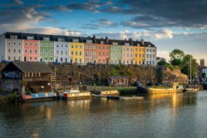Colourful pastel houses overlook boats moored along Bristols Floating Harbour in warm evening light. Reflections shimmer across the water beneath a dramatic cloudy sky.