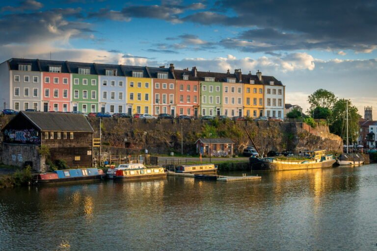 Colourful pastel houses overlook boats moored along Bristols Floating Harbour in warm evening light. Reflections shimmer across the water beneath a dramatic cloudy sky.