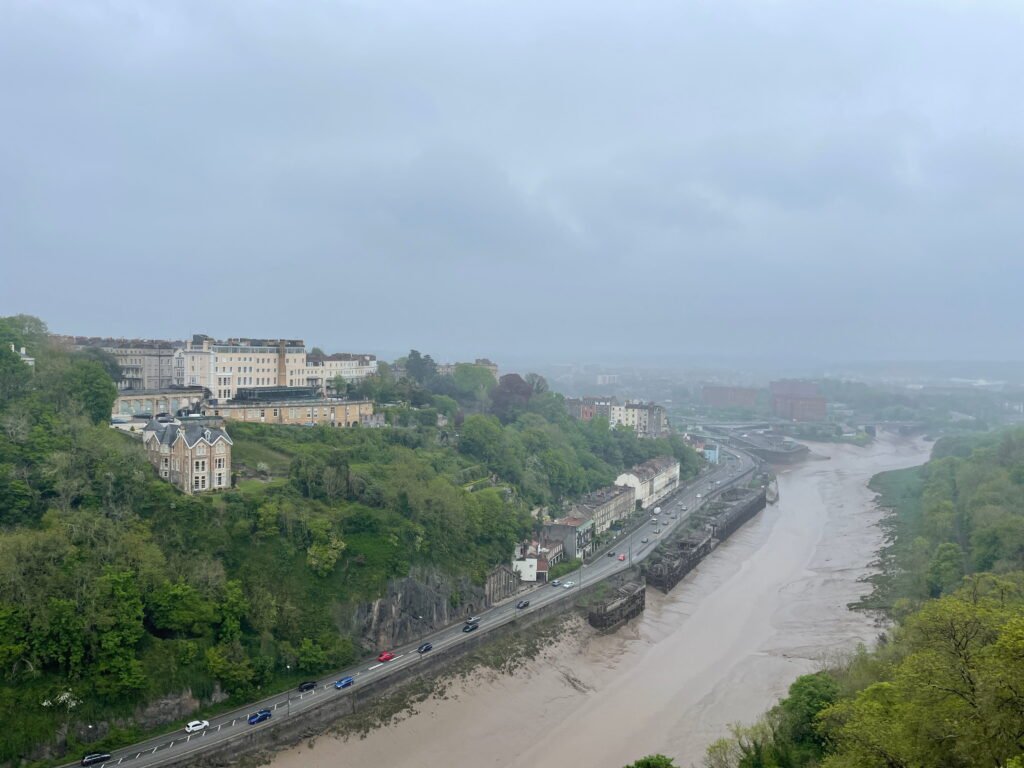 A misty view across Bristols Avon Gorge with houses and greenery covering the steep hillside above the River Avon. A road follows the muddy river below beneath a grey overcast sky.
